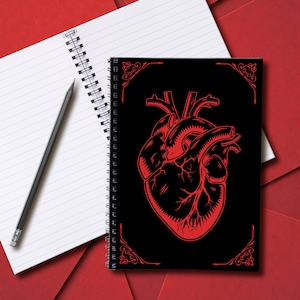 Könnte beinhalten: Ein Spiralnotizbuch mit schwarzem Einband und rotem anatomischen Herzdesign. Ein weißes liniertes Notizbuch und ein Bleistift liegen neben dem Notizbuch auf einer roten Oberfläche. Das Notizbuch hat dekorative Eckelemente.