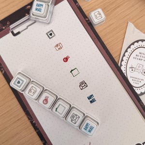Puede incluir: Un conjunto de mini sellos de botón con varios iconos temáticos de correo y las palabras "Happy Mail". Los sellos están dispuestos en un bloc de notas tipo portapapeles con un diseño de café. También se ven un pequeño cuaderno y un paquete de sellos.