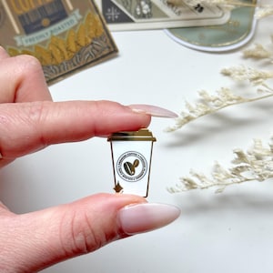 May include: Gold-tone enamel pin featuring a white coffee cup with a brown coffee bean inside. The cup has a gold rim and a gold star on the bottom. The pin is surrounded by a gold circle with the words "PREMIUM COFFEE + ROASTERS" in black text.