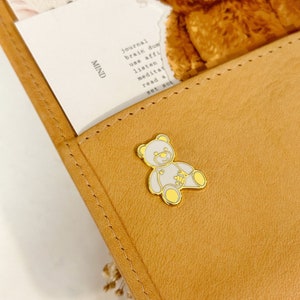 May include: A tan leather journal with a white card that says "MIND" and "journal brain dump use affirmations listen meditate read a set goal". A small white teddy bear pin is attached to the journal.