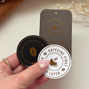 May include: Two round stickers with gold lettering on a white and black background. The black sticker says "Obsessed with Caffeine and Dreams". The white sticker says "Caffeine First, Everything Later".