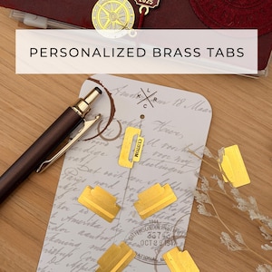 以下が含まれることがあります： 「PERSONALIZED BRASS TABS」の文字が白い長方形で表示された、金色の真鍮製パーソナライズタブのコレクション。ヴィンテージスタイルの紙の上に配置され、背景にはペンとジャーナルがあります。