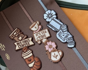 May include: Embroidered bookmarks with coffee and tea-themed designs. The bookmarks feature phrases like "But First Coffee" and "It's Tea Time." Each bookmark has a different design, including a coffee cup, a teapot, and a to-go coffee cup.