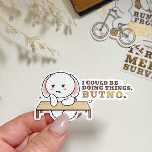 May include: A white sticker featuring a cartoon bunny with a sad expression, leaning on a table. The sticker has the text "I COULD BE DOING THINGS. BUT NO." in a gold color. Other stickers and a brown wallet are in the background.