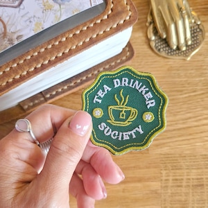 May include: A green and yellow embroidered patch with the words "TEA DRINKER SOCIETY" and a teacup graphic. The patch is held by a hand with a ring. A leather-bound journal and a decorative brass hand are in the background.