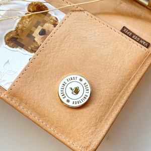 May include: A gold-coloured pin with a white background. The pin reads "Caffeine First, Adulting Later" with a coffee cup icon in the centre.