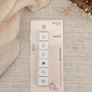 Könnte beinhalten: Ein Set aus sechs quadratischen Mini-Knopfstempeln mit der Aufschrift MOODS von KOLOUR CAFE, die schwarze Strichzeichnungen verschiedener Gesichtsausdrücke auf weißem Grund zeigen, in einer transparenten Kunststoffhalterung auf weißem Karton.