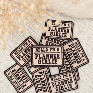 May include: A collection of rectangular embroidered patches in a light beige color with a dark brown border and dark brown text that reads "HELLO I'M A PLANNER GIRLIE" in a bold, sans-serif font, arranged in a scattered pile.