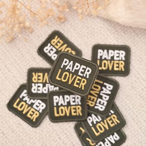 May include: A collection of square, black embroidered patches featuring the text PAPER LOVER in white and gold thread. Each patch has a stitched border and is arranged in a scattered pile, showcasing the repeating design and contrasting colors.