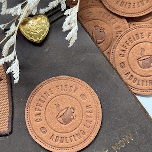 以下が含まれることがあります： コーヒーカップのアイコンを囲む円の中に「CAFFEINE FIRST ADULTING LATER」という文字が書かれた茶色の革のパッチ。パッチは「JOY THE NOW」という金色の文字が書かれた茶色の革の表面に置かれている。