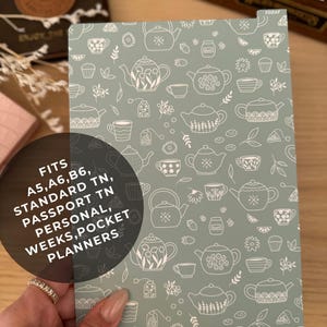 May include: A planner cover with a light green background and a white teapot pattern. The cover fits A5, A6, B6, Standard TN, Passport TN, Personal, Weeks, and Pocket planners. The text "FITS A5, A6, B6, STANDARD TN, PASSPORT TN, PERSONAL, WEEKS, POCKET PLANNERS" is visible.