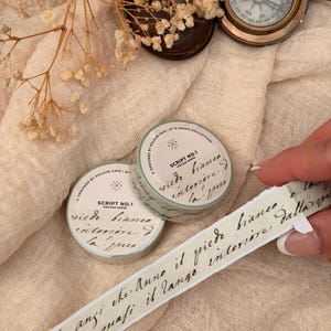 May include: Two rolls of Script No. 1 vintage series washi tape with script writing in black ink. The tape is light green with a white background. A strip of the tape is being pulled away from the roll. A compass and dried flowers are in the background.