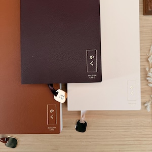 Puede incluir: Conjunto de cuadernos encuadernados en cuero en tonos marrón, burdeos y crema. Cada cuaderno presenta un detalle dorado con caracteres japoneses y una cinta marcapáginas con un charm dorado. Los cuadernos están colocados sobre una superficie de madera clara.