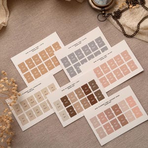 Könnte beinhalten: Mini-Planer-Tabs in verschiedenen neutralen Farben mit Monatsbeschriftungen. Die Tabs sind auf einer neutralen Oberfläche angeordnet, mit einem Vintage-Kompass und einer Kette im Hintergrund. Jeder Tab misst etwa 2 cm.