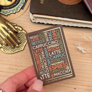 May include: A brown rectangular card with gold edging, listing coffee drinks in various colours. The card is held in a hand, with a gold-coloured hand-shaped object and a notebook in the background. The text on the card includes "Espresso", "Latte", and "Cappuccino".