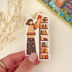 May include: A sticker featuring a cartoon illustration of a person in a floral patterned dress reaching for a book on a bookshelf filled with books. The bookshelf is brown with white shelves. The person is wearing a sweater with a floral pattern.