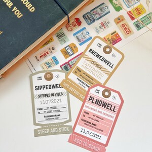 May include: A collection of decorative paper tags with travel-themed designs. The tags feature phrases like "Brewedwell," "Sippedwel," and "Plndwell," along with dates and travel-related text. A gold star and a journal are also visible.