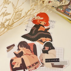 May include: Three illustrated stickers featuring girls reading books. The first sticker shows a girl with red hair sitting on a chair with the text "Cozy" above her. The second sticker shows a girl with brown hair sitting in a chair with a blanket. The third sticker shows a girl with black hair sitting in a chair with a blanket and the text "Bookworm" below her.