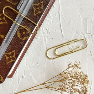 Puede incluir: Un cuaderno de cuero marr&oacute;n con un clip de papel dorado sujetando un bol&iacute;grafo. Un clip de papel dorado est&aacute; sobre la superficie blanca junto al cuaderno. Un ramo de flores blancas secas est&aacute; sobre la superficie blanca.
