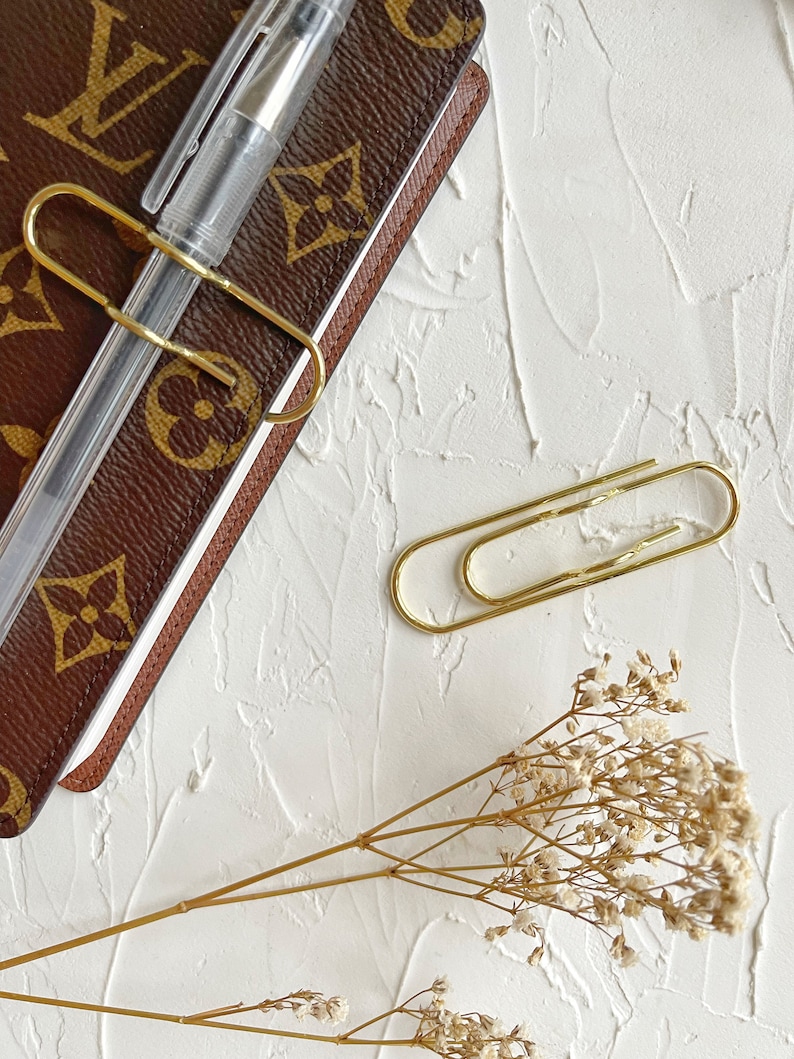 Puede incluir: Un cuaderno de cuero marr&oacute;n con un clip de papel dorado sujetando un bol&iacute;grafo. Un clip de papel dorado est&aacute; sobre la superficie blanca junto al cuaderno. Un ramo de flores blancas secas est&aacute; sobre la superficie blanca.