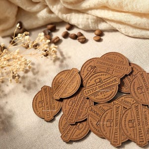Peut inclure: Plusieurs étiquettes rondes en similicuir marron, portant l'inscription en relief "LITTLE NOTEBOOK BIG STORIES". Elles sont posées sur un tissu beige, avec des grains de café et une brindille de fleurs séchées en arrière-plan.