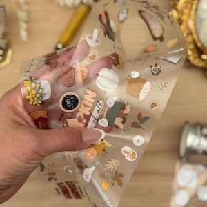 May include: A roll of clear washi tape with autumn-themed illustrations. The tape features images of pumpkins, pears, mushrooms, and text. The tape is held by a hand, with other decorative items in the background.