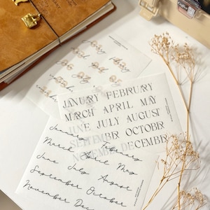 May include: Two sheets of clear stickers with the months of the year printed in black and white. The top sheet has the months printed in a sans-serif font, while the bottom sheet has the months printed in cursive.