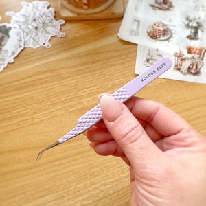 May include: A pair of purple tweezers with a diamond pattern on the handle. The tweezers have a slanted tip and are labeled "KOLOUR CAFE".