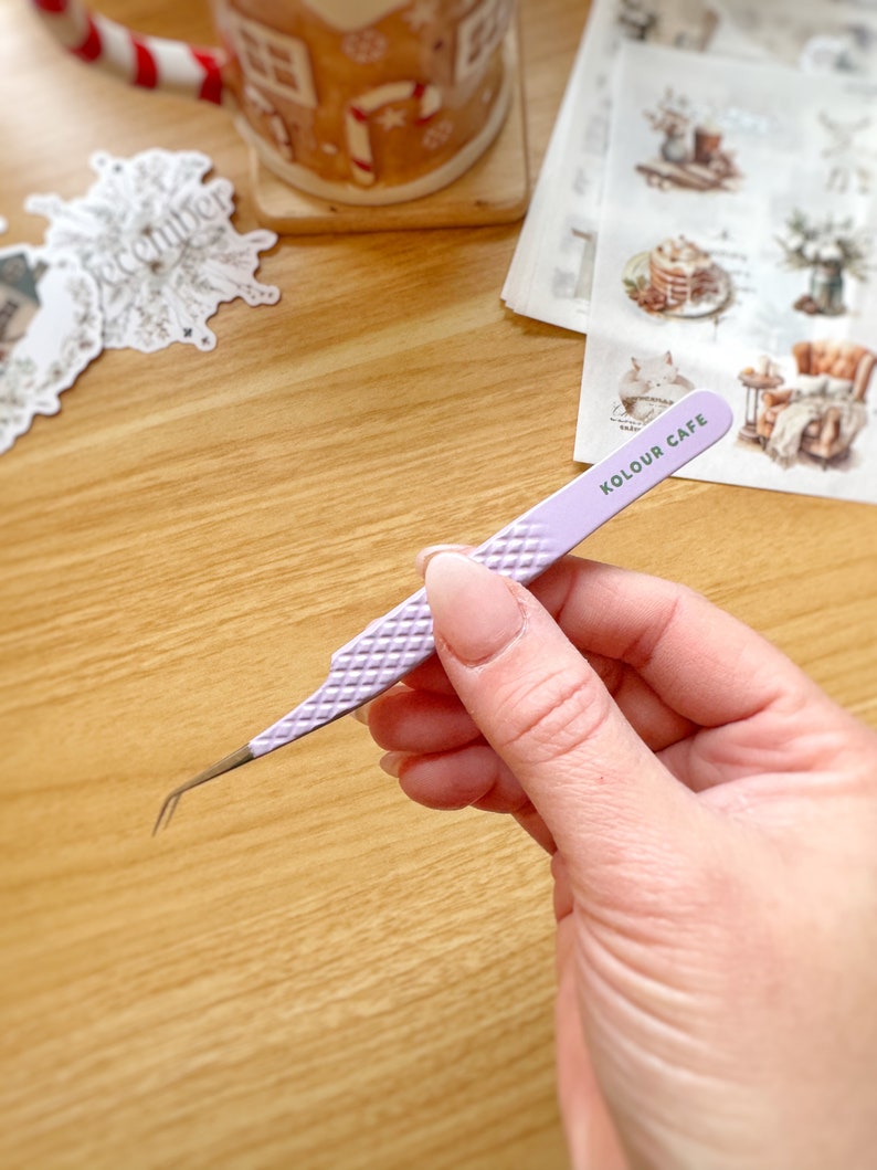 May include: A pair of purple tweezers with a diamond pattern on the handle. The tweezers have a slanted tip and are labeled "KOLOUR CAFE".