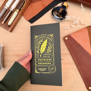 Könnte beinhalten: Ein dunkelgraues Notizbuch mit goldfarbenen Buchstaben, auf dem "Places, Moments & Memories" und "Made for Travelers Dreamers" steht. Das Notizbuch wird von einer Person gehalten. Weitere Artikel sind Stifte, Tinte und Lederwaren.