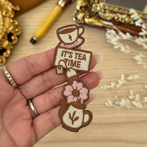 May include: Embroidered patch featuring a tea cup, a sign that reads "IT'S TEA TIME", a pink flower, and a teapot. The patch is brown with pink and white accents. The patch is held in a hand.