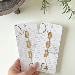 Peut inclure: Deux paires de boucles d'oreilles sur cartes. Chaque carte présente un design vintage avec une écriture manuscrite et un logo de café. Les boucles d'oreilles sont dorées, de forme rectangulaire, avec une petite étiquette "WEEKLY".