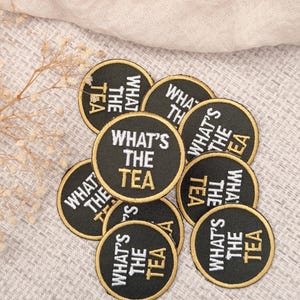 May include: A collection of circular black embroidered patches featuring a gold border. Each patch displays the text "WHAT'S THE TEA" in white and gold thread, with "WHAT'S THE" in white and "TEA" in gold, arranged in a stacked, centered layout.