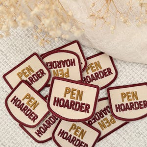 May include: A collection of shield-shaped embroidered patches featuring a cream-colored background with a dark red border. Each patch displays the text PEN HOARDER in stacked lines, with PEN stitched in gold thread and HOARDER in dark red thread.