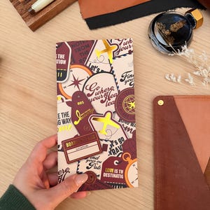 Puede incluir: Un diario de viaje burdeos y crema con detalles dorados, sostenido por una mano. El diario presenta frases como "Go where your heart leads" y "Boarding Pass". Otros artículos incluyen bolígrafos, tinta y artículos de cuero.