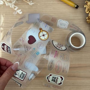 Puede incluir: Pegatinas transparentes con temática de viajes con sellos de destino, una brújula y los textos "Let's go TRAVEL" y "TAKE THE LONG WAY home". También se ve un rollo de cinta adhesiva transparente. Sobre una superficie de madera clara.