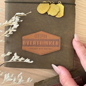 May include: A brown leather journal with a leather patch that reads "CERTIFIED OVERTHINKER - PROCEED WITH CAUTION." The journal has a black elastic closure and gold charms. A sprig of dried flowers is on the side.