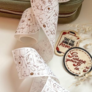Puede incluir: Un rollo de cinta washi blanca con un patrón marrón y blanco de tazas de café, accesorios de viaje y otros artículos cotidianos. La cinta está desenrollada y muestra el patrón que se repite. La cinta está sobre una superficie blanca.