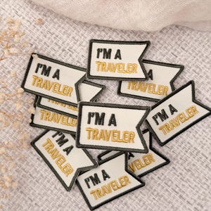 May include: A collection of small, flag-shaped embroidered patches with a white background and a black border. Each patch features the text "I'M A TRAVELER" embroidered in black for the first line and gold-colored thread for the second line.