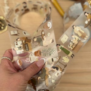 May include: Clear decorative tape featuring illustrations of people in a cafe setting, with coffee cups, plants, and cafe signage. The tape is held in a hand with a ring. The tape is on a wooden surface.