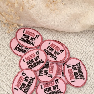 May include: A collection of oval-shaped embroidered patches in light pink with a dark pink border. Each patch features the text "I NEED IT FOR MY JOURNAL" embroidered in bold black capital letters, centered within the oval design.