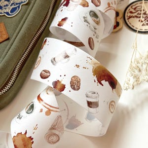 May include: White washi tape with a coffee theme. The tape features illustrations of coffee cups, coffee beans, and coffee stains.