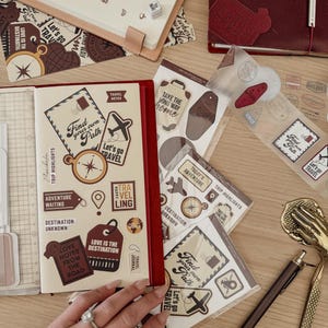 Może przedstawiać: Kolekcja artykułów papierniczych o tematyce podróżniczej, w tym naklejki, notatniki i stemple. Naklejki zawierają frazy takie jak "Find your own Path" i "Let's go TRAVEL". Notatniki są w kolorze bordowym i kremowym.