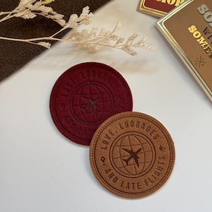 May include: Two round leather patches with a globe and airplane design. The text on the patches reads "Love, Luggages and Late Flights".