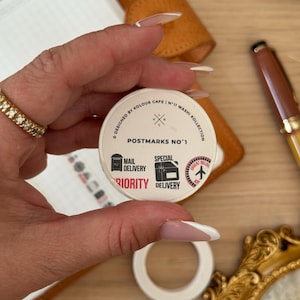 以下が含まれることがあります： 郵便をテーマにしたマスキングテープのロール。テープのラベルには「POSTMARKS NO.1」と書かれており、郵便配達と優先配達のシンボルが描かれています。テープは白で円形のデザインです。