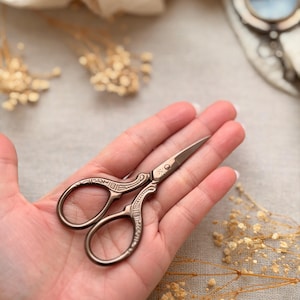 May include: Antique bronze-colored embroidery scissors with ornate detailing. The scissors are open and resting on a hand, with a small cross design near the pivot. The background includes dried flowers and a magnifying glass.
