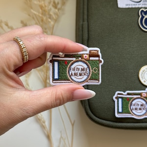 Pode incluir: Etiqueta de ferro bordada verde e branca de uma câmera com o texto "Every mile a memory".