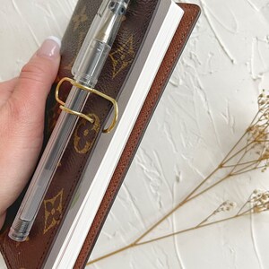 Puede incluir: Una libreta de cuero marr&oacute;n con un portaplumas dorado y una pluma transparente. La libreta tiene una cubierta con un patr&oacute;n marr&oacute;n y beige.