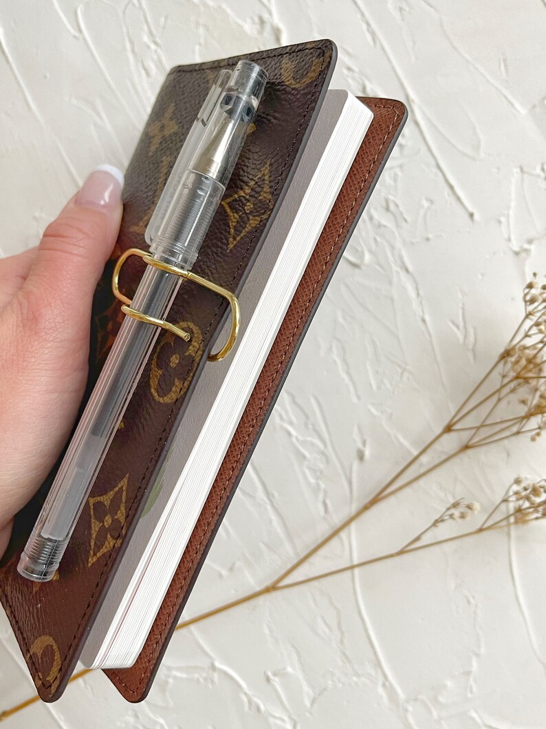 Puede incluir: Una libreta de cuero marr&oacute;n con un portaplumas dorado y una pluma transparente. La libreta tiene una cubierta con un patr&oacute;n marr&oacute;n y beige.