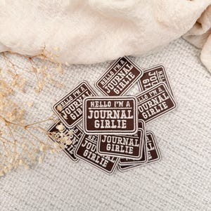 May include: A collection of rectangular embroidered patches in dark brown with white borders and white text. Each patch features the phrase HELLO I'M A JOURNAL GIRLIE in a bold, sans-serif font, arranged in a stacked layout on a textured surface.